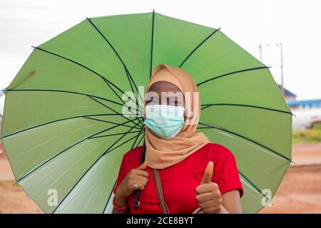 Junge schöne Frau in Hijab trägt einen Regenschirm und trägt Eine medizinische Maske Stockfoto