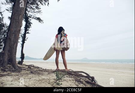 Rückansicht von unerkennbarer junger Surferin im Sommer-Outfit Wandern am Sandstrand und Surfbrett gegen ruhiges Blau tragen Blick auf das Meer Stockfoto