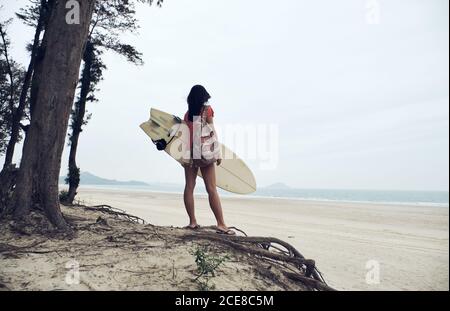 Rückansicht von unerkennbarer junger Surferin im Sommer-Outfit Wandern am Sandstrand und Surfbrett gegen ruhiges Blau tragen Blick auf das Meer Stockfoto