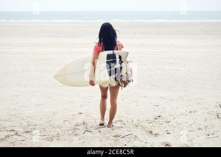 Volle Länge Rückansicht von nicht erkennbaren jungen weiblichen Surfer in Sommer Outfit Wandern am Sandstrand und tragen Surfbrett gegen Ruhiges blaues Meer, das weg schaut Stockfoto