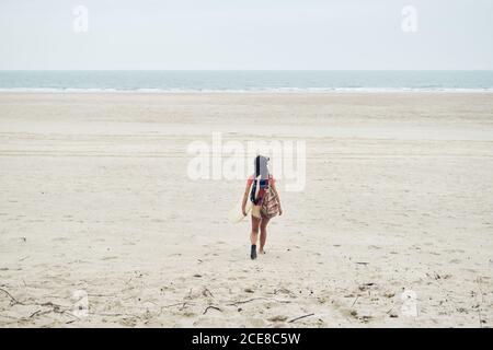 Volle Länge Rückansicht von nicht erkennbaren jungen weiblichen Surfer in Sommer Outfit Wandern am Sandstrand und tragen Surfbrett gegen Ruhiges blaues Meer, das weg schaut Stockfoto