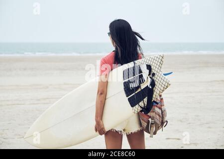 Rückansicht von unerkennbarer junger Surferin im Sommer-Outfit Wandern am Sandstrand und Surfbrett gegen ruhiges Blau tragen Blick auf das Meer Stockfoto