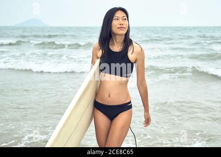 Junge asiatische Surferin zu Fuß auf Sandstrand und tragen Surfbrett gegen ruhiges blaues Meer, das wegschaut Stockfoto