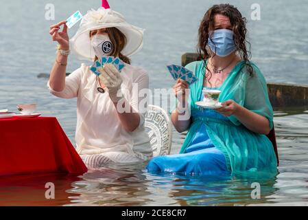 Southend on Sea, Essex, Großbritannien. August 2020. Der lokale Zweig der Extinction Rebellion hat ein einzigartiges Ereignis inszeniert, um die steigenden Gezeiten durch den Klimawandel zu symbolisieren, indem er einen Tisch im Meer für den Nachmittagstee aufstellt, während die Flut im Badeort eintrifft. Gekleidet in edwardianische Kleidung ist die Idee, die Band zu repräsentieren, die als Titanic sinkt. Zwei Frauen tragen Gesichtsmasken, trinken Tee und Spielkarten Stockfoto