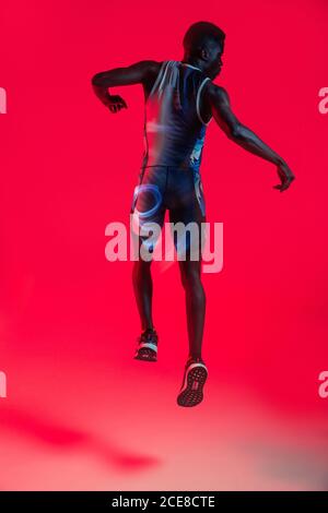 Ganzkörper-Rückansicht des professionellen jungen afroamerikanischen Sportlers Im sportiven Outfit springen im Studio mit roter Neonbeleuchtung Stockfoto