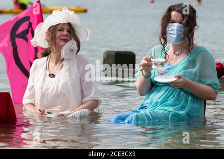 Southend on Sea, Essex, Großbritannien. August 2020. Der lokale Zweig der Extinction Rebellion hat ein einzigartiges Ereignis inszeniert, um die steigenden Gezeiten durch den Klimawandel zu symbolisieren, indem er einen Tisch im Meer für den Nachmittagstee aufstellt, während die Flut im Badeort eintrifft. Gekleidet in edwardianische Kleidung ist die Idee, die Band zu repräsentieren, die als Titanic sinkt. Zwei Frauen, die Tee trinken, sitzen im Meerwasser Stockfoto