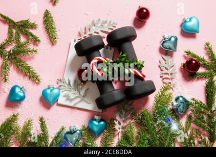 Flache Lay-Ansicht von schwarzen Hanteln mit weißem Spiralheft mit Weihnachtsornamenten auf rosa Hintergrund. Gesunde Lebensweise während des Weihnachtsfests. Stockfoto