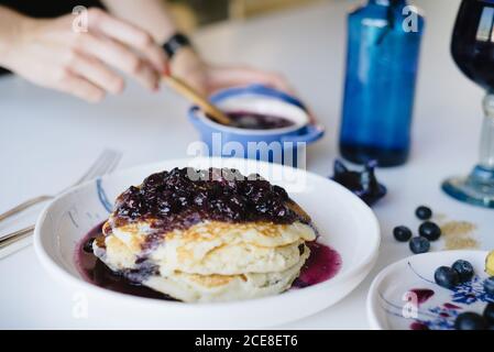 Von oben der Ernte anonyme Frau, die yummy Heidelbeermarmelade nimmt Mit Löffel, während Sie Pfannkuchen zum Frühstück zu Hause servieren Stockfoto