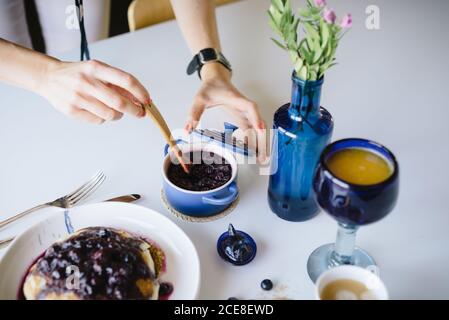 Von oben der Ernte anonyme Frau, die yummy Heidelbeermarmelade nimmt Mit Löffel, während Sie Pfannkuchen zum Frühstück zu Hause servieren Stockfoto