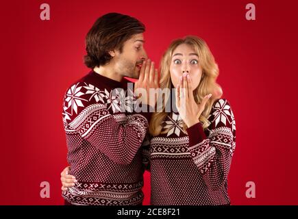 Schockiert junge Dame in Weihnachten Pullover und Freund flüstert Geheimnis In ihr Ohr auf rotem Hintergrund Stockfoto