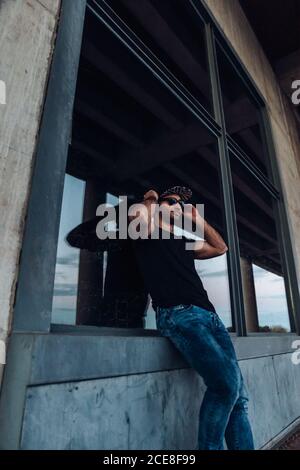 Niedriger Winkel des selbstbewussten jungen bärtigen männlichen trägt schwarzes Hemd Und Jeans mit Mütze und Sonnenbrille, die beim Anlehnen wegschauen Gegen Fenster des verwitterten Steingebäudes Stockfoto