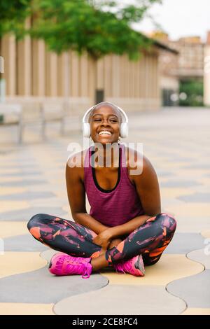 Positive afroamerikanische Sportlerin in Sportkleidung und Kopfhörern Sitzend lehnte sich nach vorne mit gekreuzten Beinen auf dem Damm neben dem Gebäude In der Stadt und Blick auf die Kamera, während Sie Musik hören Stockfoto