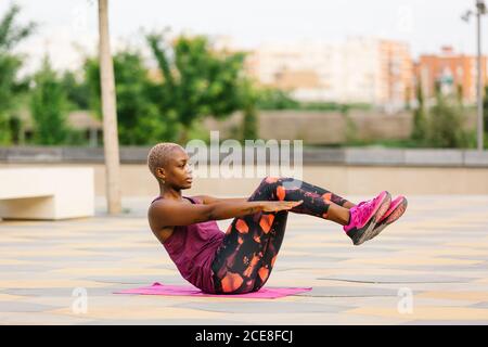 Seitenansicht einer afroamerikanischen FIT-Frau in farbenfroher aktiver Kleidung, die in Ardha Navasana auf einem kleinen Handtuch sitzt, während sie bei Tageslicht auf dem Bürgersteig in der Stadt in der Nähe von Gebäuden Yoga praktiziert Stockfoto