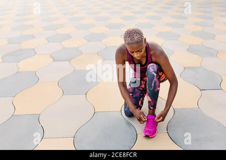 Schwarze Sportlerin in farbenfroher aktiver Kleidung, die Schnürsenkel auf Schuhen bindet, während sie im Sommer auf dem Bürgersteig in der Stadt in der Nähe von Gebäuden unter heiterem Himmel hockt Stockfoto