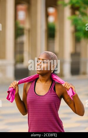 Junge afroamerikanische Sportlerin mit gefärbten Haaren im Sport Kleidung mit rosa Baumwolltuch auf Schultern stehend mit offenen Mund auf Bürgersteig in der Stadt in der Nähe von Gebäude und Blick weg Stockfoto