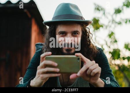 Inhalt männlichen Hipster mit langen Haaren und Rucksack mit einem Telefon und Blick auf die Kamera während der Sommerferien Stockfoto