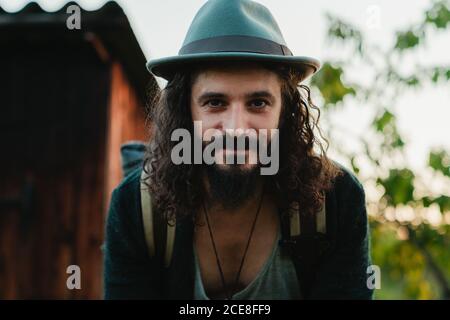 Inhalt männliche Hipster mit langen Haaren und Rucksack stehen in Landschaft und Blick auf die Kamera während der Sommerferien Stockfoto