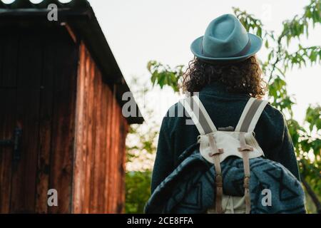 Von hinten Blick auf anonymen Mann Hipster mit langen Haaren Und Rucksack, der auf dem Land neben einer Holzhütte steht Während der Sommerferien Stockfoto