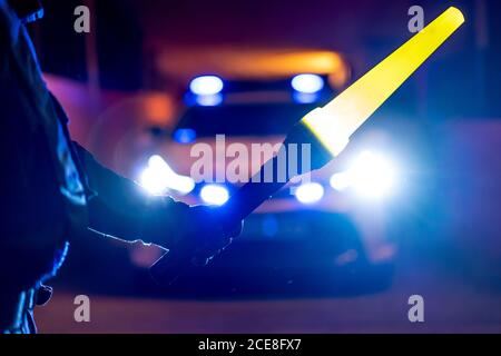 Rückansicht des Getreidepoliziers stehend mit beleuchtetem Verkehr Zauberstab vor dem Streifenwagen mit Blinklicht an Nacht Stockfoto