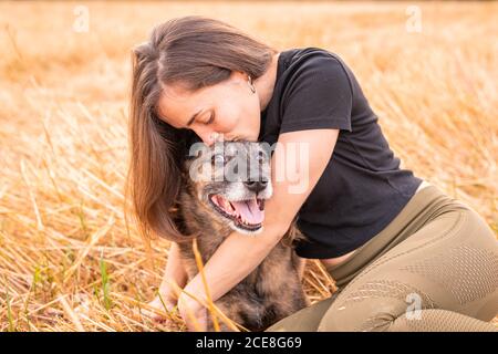 Weibchen mit geschlossenen Augen kuscheln sanft reinrassigen Hund mit glatt Mantel, während er auf dem Feld mit verblassenem Gras in der Nähe des Waldes sitzt In der Landschaft unter dem Himmel mit Wolken Stockfoto