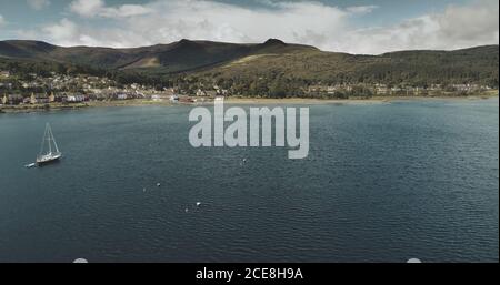 Atlantischer Ozean Golf, Segelboote Luftzoom in Brodick Bay aufgenommen. Schottische Landschaft der Hafenstadt. Häuser, Ferienhäuser, Resort am Ufer des Golfs gegen grüne Länder und Berge Blick Stockfoto