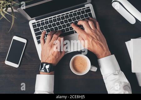 Draufsicht auf die Hände eines Geschäftsreisenden, der die Tastatur eines Laptops mit einer Tasse Kaffee auf dem Schreibtisch tippt, Ansicht des Arbeitsbereichs von oben Stockfoto