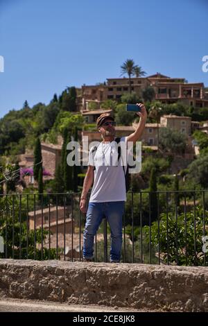 Fröhlicher Hipster bärtiger männlicher Tourist in stylischer Sonnenbrille und Mütze Selfie auf dem Smartphone beim Besuch der historischen Siedlung im Sommer Reise Stockfoto