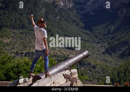 Rückansicht von aufgeregt bärtigen männlichen Touristen in stilvoller Kleidung Stehen mit Arm auf alten Kanonen gegen Berge während angehoben Sommerurlaub Stockfoto