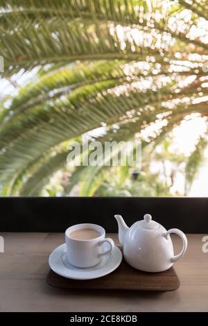 Weiße Teekanne mit Tasse Masala Tee auf dem Tisch in der Nähe einer Palme und wunderbares Licht mit Sonnenstrahlen, vertikal Stockfoto