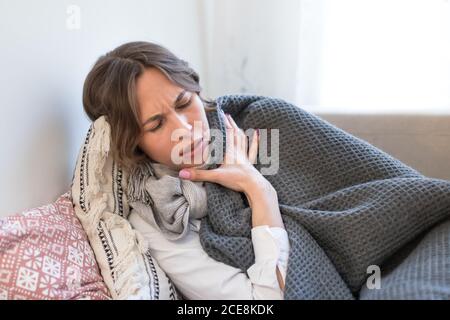 Kranke junge Frau liegt und zittert, eingewickelt in Decke und Schal, versucht sich zu wärmen, mit Grippesymptomen Stockfoto