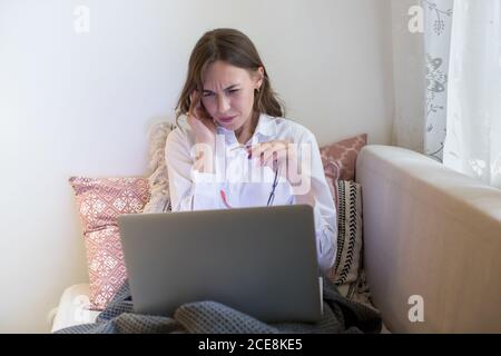 Kopfschmerzen. Müde Weibchen hat Kopfschmerzen, nachdem sie am Laptop gearbeitet hat, ihren Tempel berührt, eine Brille hält, versucht sich zu konzentrieren Stockfoto