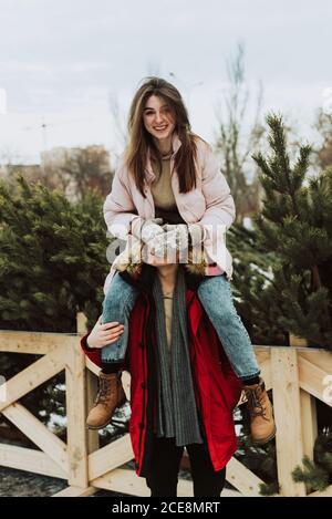 Eine junge Frau sitzt auf den Schultern eines Mannes vor dem Hintergrund von Weihnachtsbäumen, schließt die Augen ihres Mannes und hat Spaß. Winterporträt von Liebenden. Stockfoto