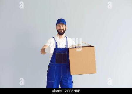 Lieferer hält einen Karton und zeigt Daumen nach oben auf einem grauen Wandhintergrund. Stockfoto