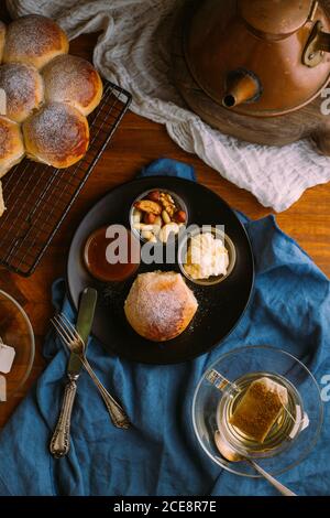 Draufsicht auf Keramikplatte mit frischem Brötchen und klein Schüsseln mit Mischung von Nüssen in der Nähe von Marmelade und Butter schließen Zu einer Tasse grünem Tee auf einem zerknitterten Handtuch Stockfoto