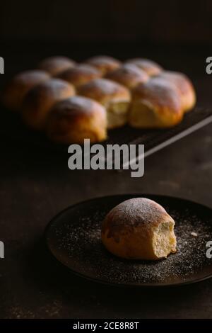 Von oben köstliche frische runde Brötchen mit weicher Textur Und goldene Oberfläche mit Puderzucker verziert auf Kühlregal Und Tisch auf schwarzem verschwommenem Hintergrund Stockfoto