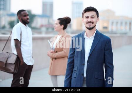 Porträt eines lächelnden jungen bärtigen Geschäftsmann in formeller Jacke stehend Gegen multiethnische Kollegen auf der Straße Stockfoto