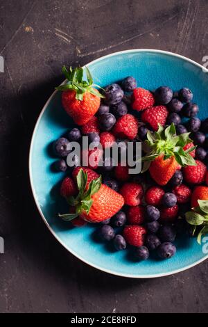 Draufsicht auf blaue Schale mit leckeren reifen Erdbeeren und Heidelbeeren mit Himbeeren auf schwarzem Hintergrund angeordnet Stockfoto