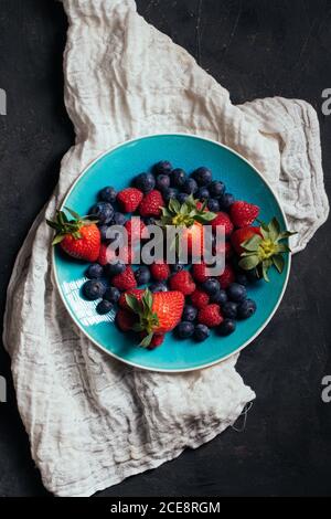 Draufsicht auf blaue Schale mit leckeren reifen Erdbeeren und Heidelbeeren mit Himbeeren auf grauem Tuch auf schwarzem Hintergrund angeordnet Stockfoto