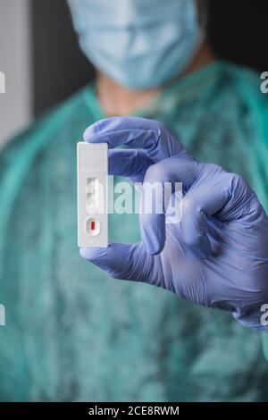 Nicht erkennbarer Arzt mit Gesichtsmaske in medizinischen Handschuhen mit Ergebnissen Des schnellen COVID 19-Tests auf grauem Hintergrund im Krankenhaus Stockfoto
