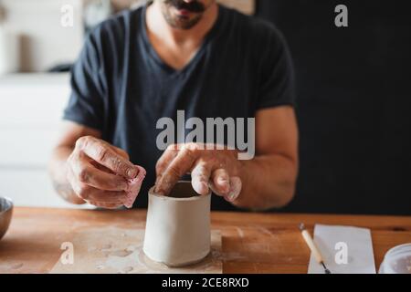 Nicht erkennbarer Keramiker mit Ton und die Schaffung Tasse während des Sitzens Tisch und Arbeiten in der Werkstatt Stockfoto