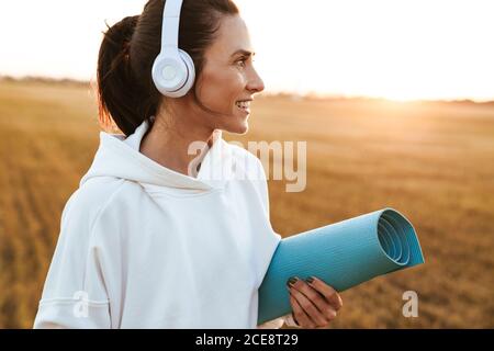Bild von lächelnder, schöner Sportlerin mit Kopfhörern, während sie Yoga hält Matte auf goldenem Feld Stockfoto