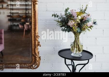 Schöner Frühlingsstrauß mit verschiedenen bunten blühenden Blumen einschließlich rosa Pfingstrosen und blaue Seeteumchen Blumen mit Mimosenzweigen angeordnet Auf Glasvase auf Tisch in der Nähe der weißen Wand Stockfoto
