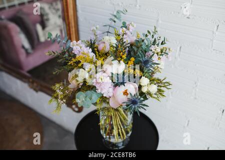 Hoher Winkel von schönen Frühling Bouquet mit verschiedenen bunten blühenden Blumen einschließlich rosa Pfingstrosen und blauen Meer Stechpalme Blumen mit mimosa Zweige auf Glasvase auf Tisch in der Nähe weiß angeordnet Wand Stockfoto