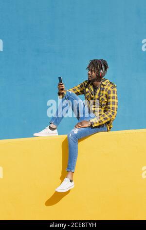Junger schwarzer Mann sitzt auf einer gelben Wand mit Kopfhörern und berät sein Smartphone mit einer blauen Wand im Hintergrund, Technologie- und Lifestyle-Konzept, Kopierraum für Text Stockfoto