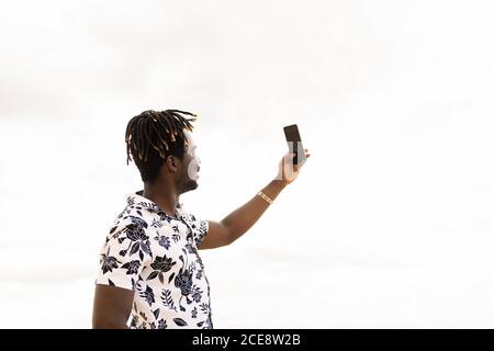 Stilvoller junger schwarzer Mann, der mit seinem Handy ein Selfie-Foto mit dem Himmel im Hintergrund macht, Technologie- und Lifestyle-Konzept, Kopierraum für tex Stockfoto