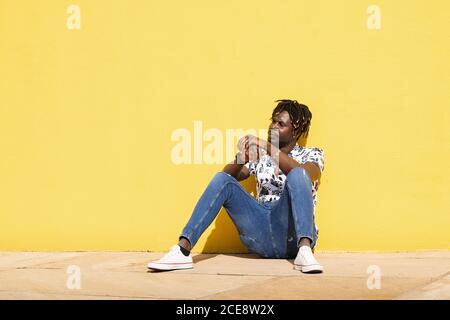 Entspannter junger afrikanischer Mann sitzt auf dem Boden gegen eine gelbe Wand, Lifestyle-Konzept, Kopierraum für Text Stockfoto