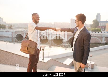 Positive junge multi-ethnische Business-Studenten auf Treppe in der Stadt stehen Und die Faust stoßen, während sie sich gegenseitig grüßen Stockfoto