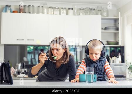 Frau in der Küche essen und beobachten elektronische Tablette. Das Kind sitzt auf dem Tisch in Kopfhörern und schaut auf das Mobiltelefon Stockfoto