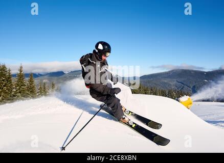 Konzentriertes Mann-Skifahrer-Training auf künstlichem Tiefhang mit frischem Neuschnee an sonnigen Tagen. Elektrische Schneekanone Maschine in Aktion auf Hintergrund. Blauer Himmel und malerische bewaldete Berge im Hintergrund Stockfoto