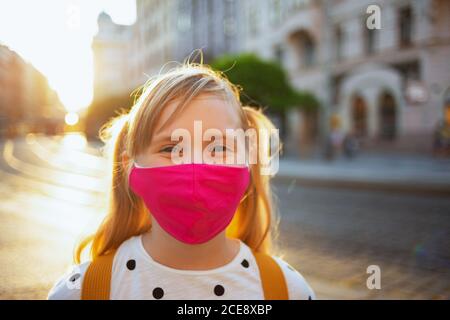 Leben während der covid-19 Pandemie. Portrait von modernen Mädchen in weißen Polka dot Bluse mit rosa Maske und gelben Rucksack gehen zur Schule im Freien. Stockfoto